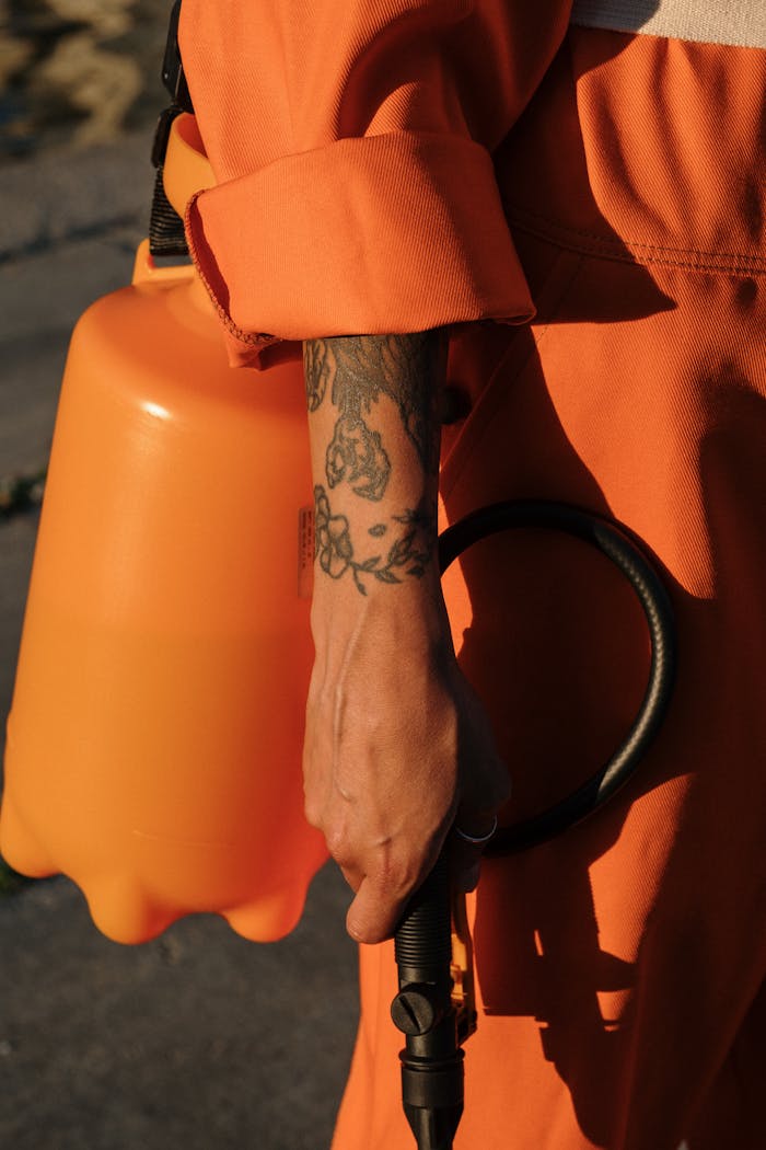 hero-01 Person in orange coveralls holding a plastic sprayer, focus on tattooed arm.