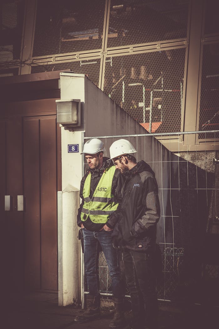 services-01 Two construction workers in safety gear standing outdoors at a building site, focused on work.